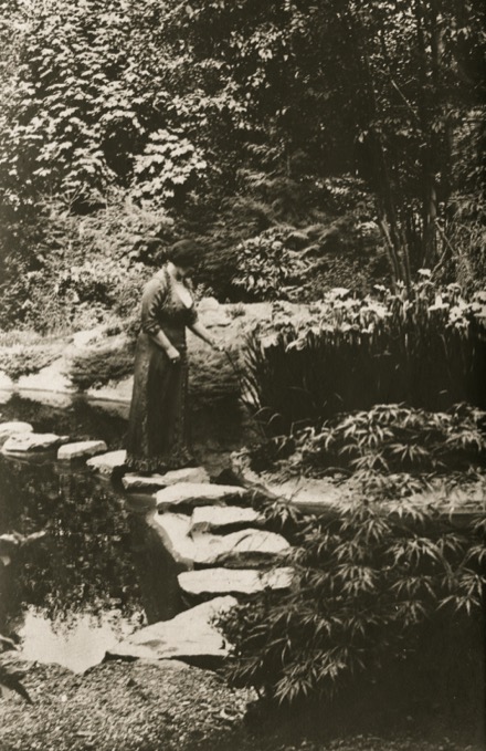 Sur cette photographie en noir et blanc, Jennie Butchart se promène dans son jardin. Elle se tient sur l’un des pavés du sentier qui traverse le ruisseau. À l’arrière-plan, on aperçoit des arbres feuillus. Au premier plan, des arbustes de différentes tailles bordent le sentier et le ruisseau.